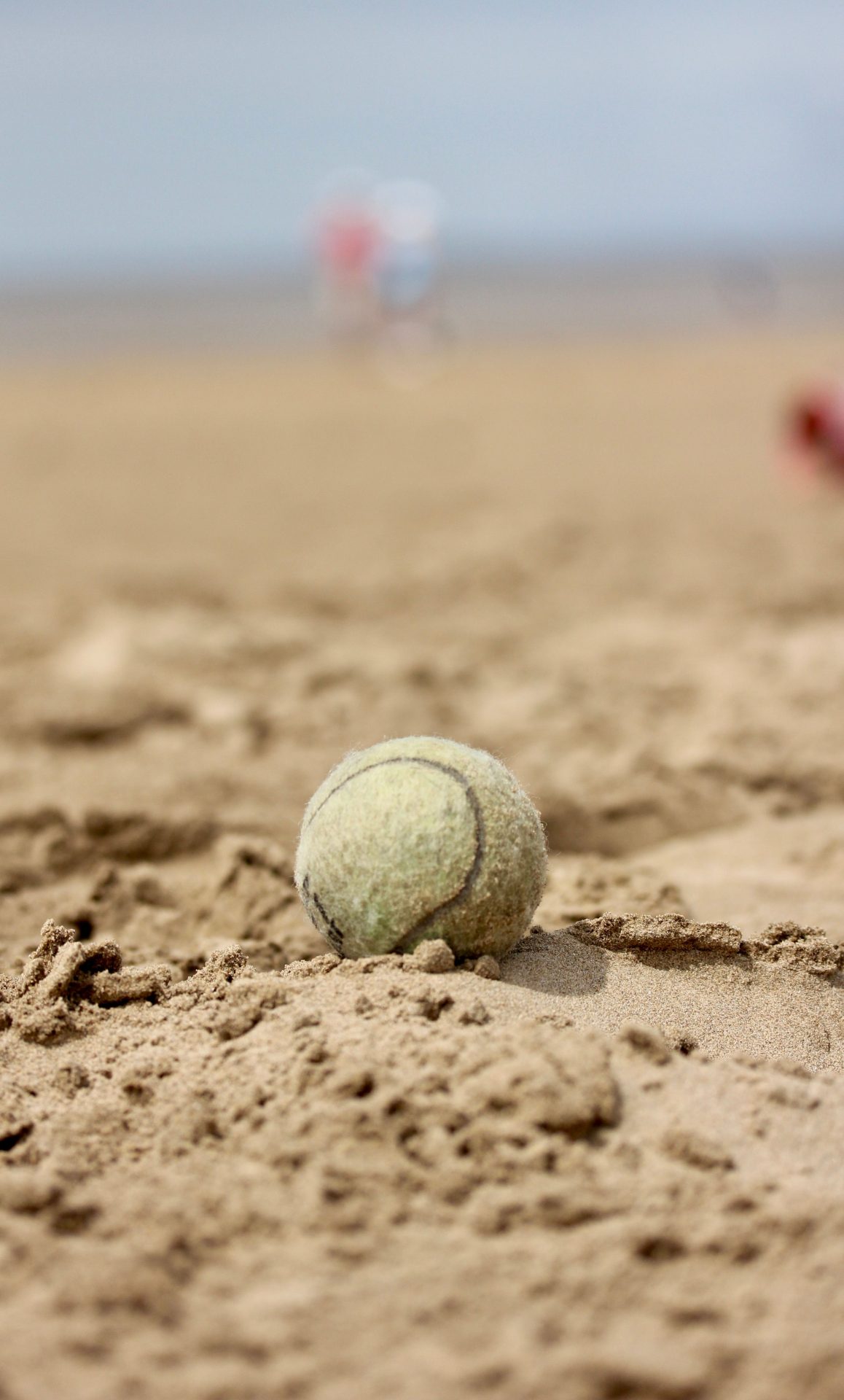 A lone tennis ball resting on a sandy beach with blurred figures in the background.